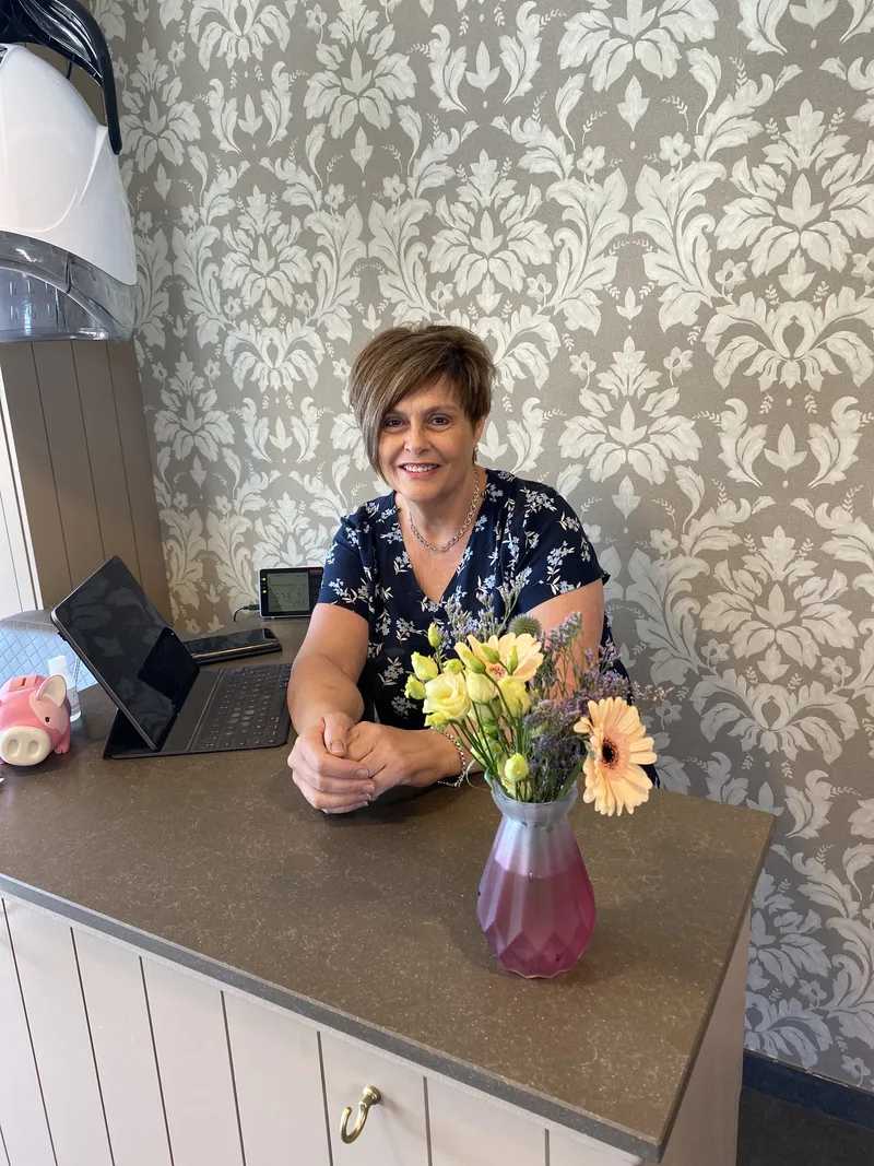 Ann achter de ontvangstbalie van haar salon, met een tablet en een vaas bloemen
